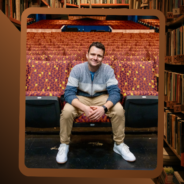 A man in a sweater sits on a red theater seat with patterned upholstery in an empty auditorium, surrounded by bookshelves.